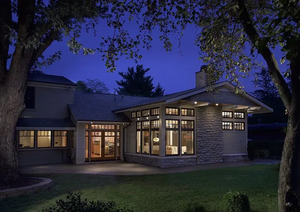 Night view of a house with many windows, illuminated and framed by trees on a green lawn.