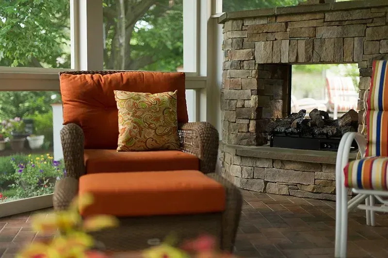Cozy porch with orange chair and ottoman, a stone fireplace, and a garden view.