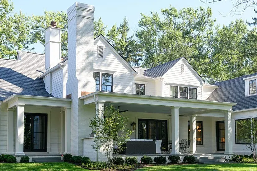 White house with a covered porch, black trim, and chimney, set in a green yard with trees.