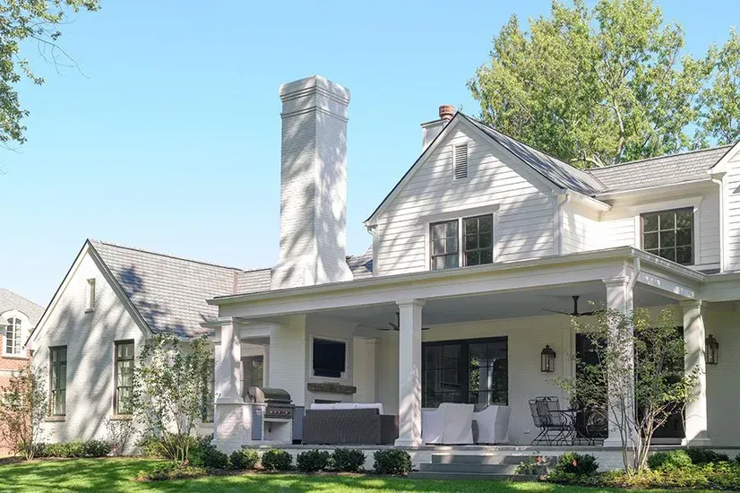 White house with a covered porch, chimney, and green lawn under a blue sky.