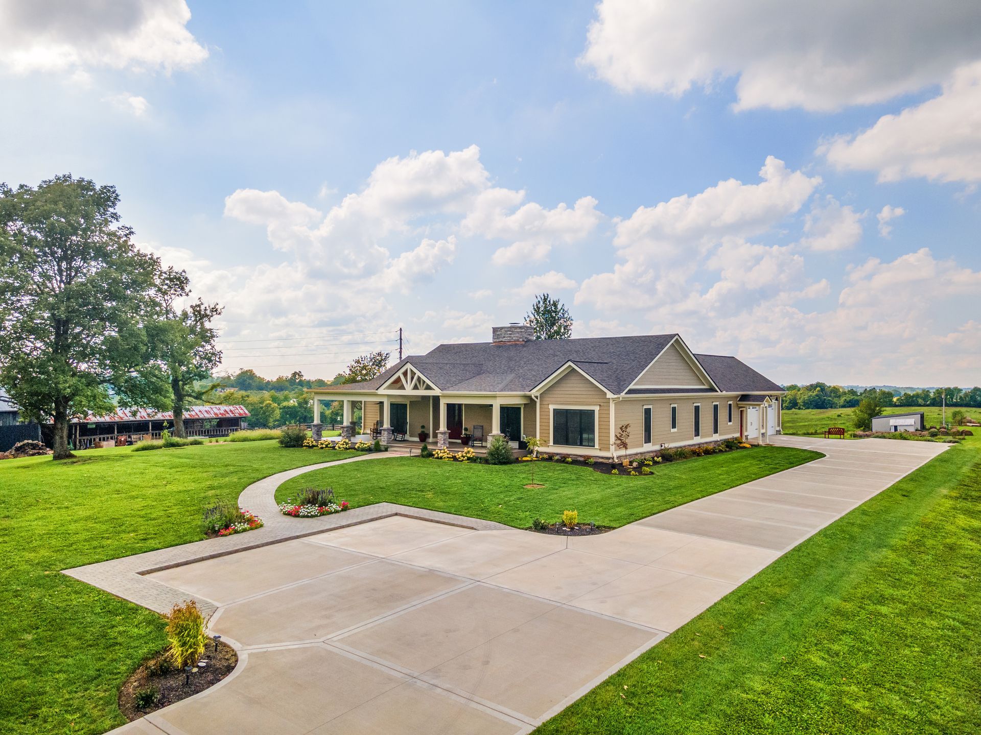 Beige ranch-style house with a long driveway, lush green lawn, and blue sky with clouds.