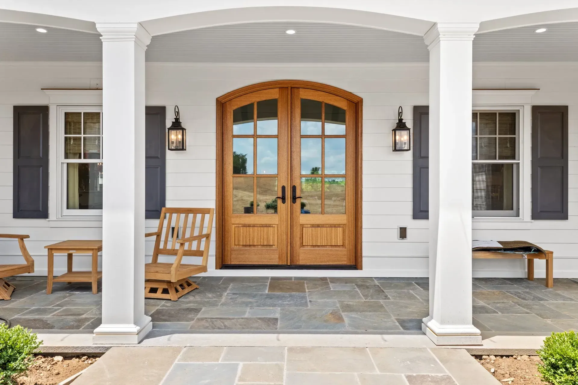 White porch with a wooden door, flanked by windows with black shutters, blue-gray stone floor.