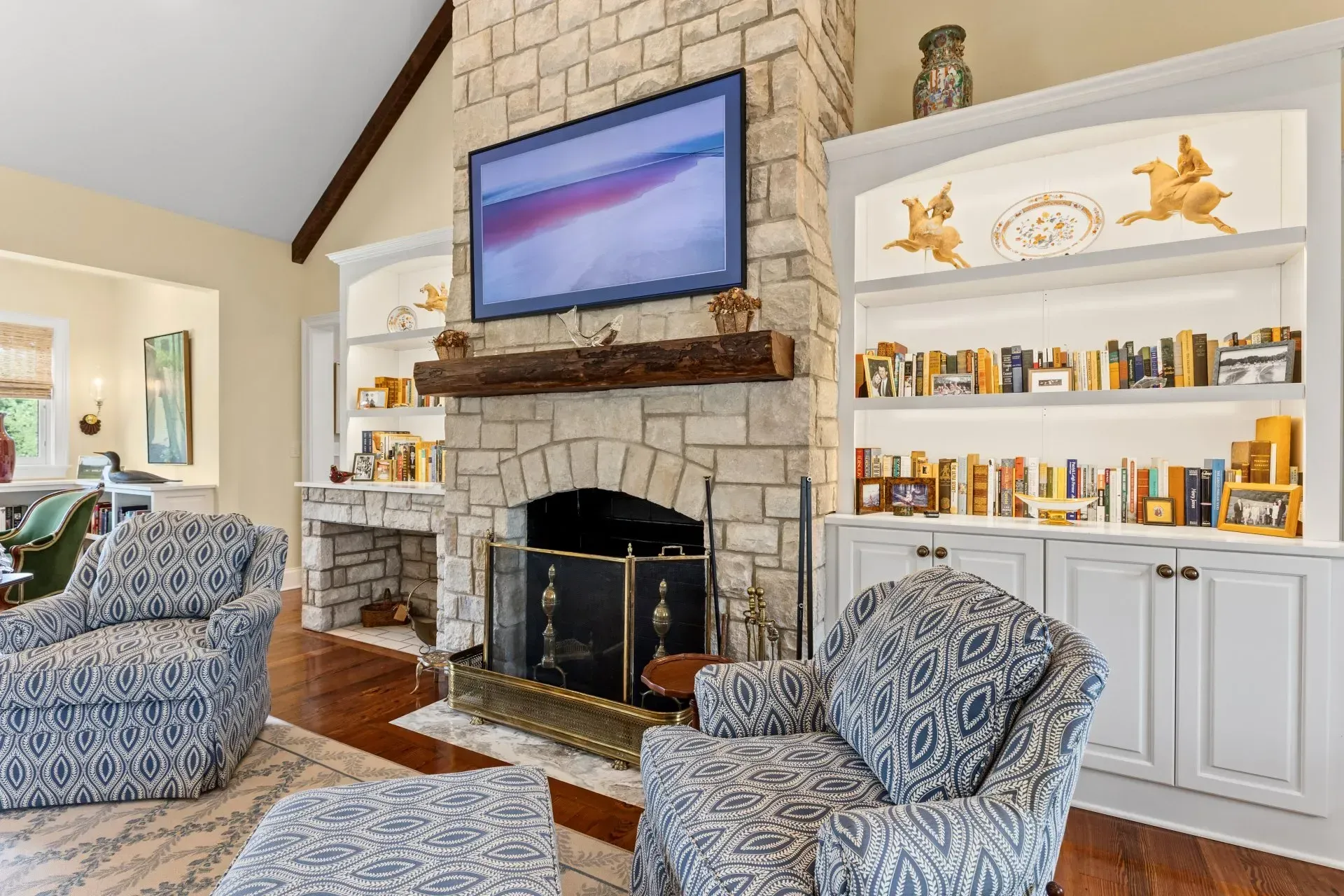 Cozy living room with stone fireplace, built-in bookshelves, TV, two patterned armchairs, and wooden floors.