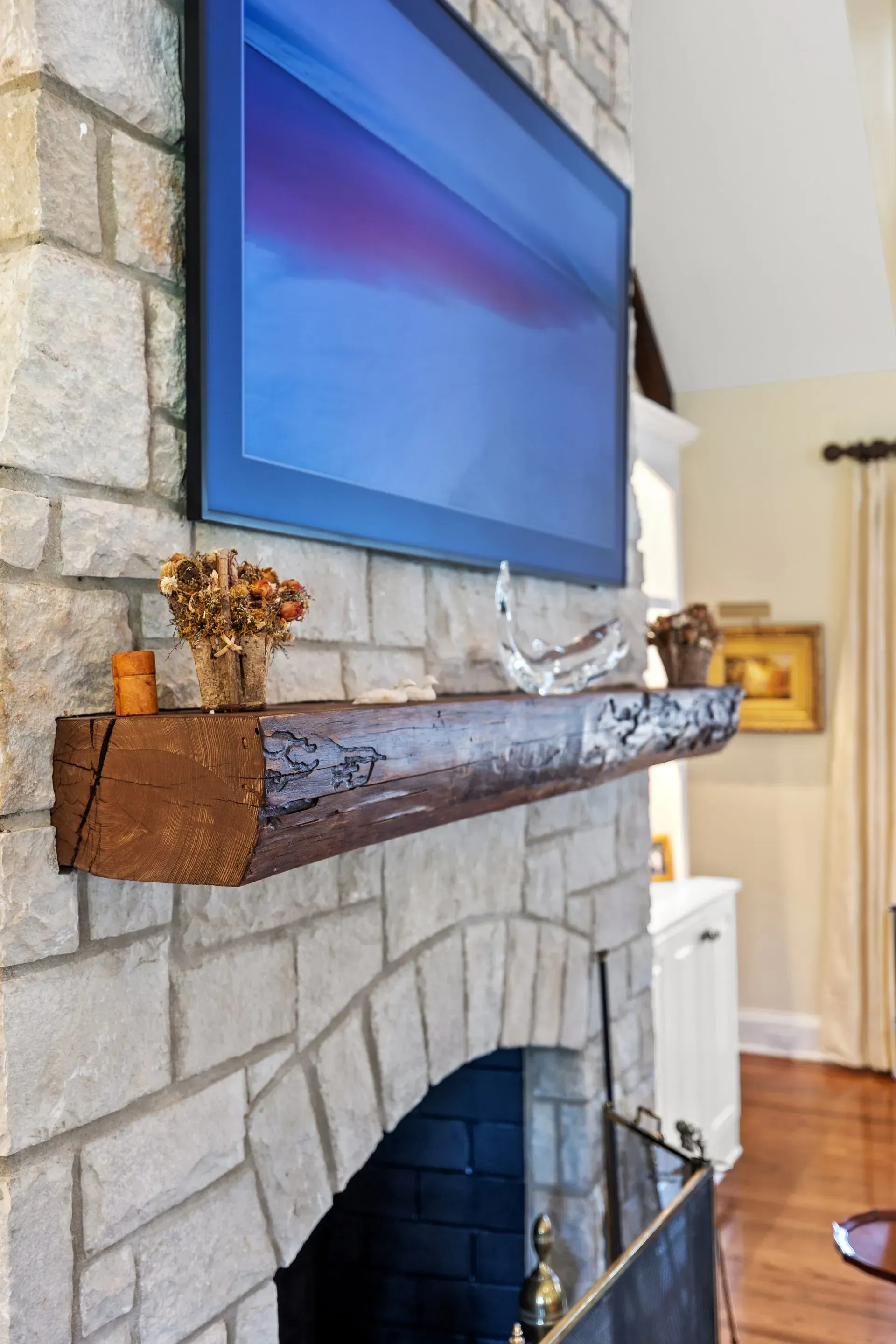 Fireplace with stone facade, dark wood mantel, and large TV above. Dried flowers and decor adorn the mantel.