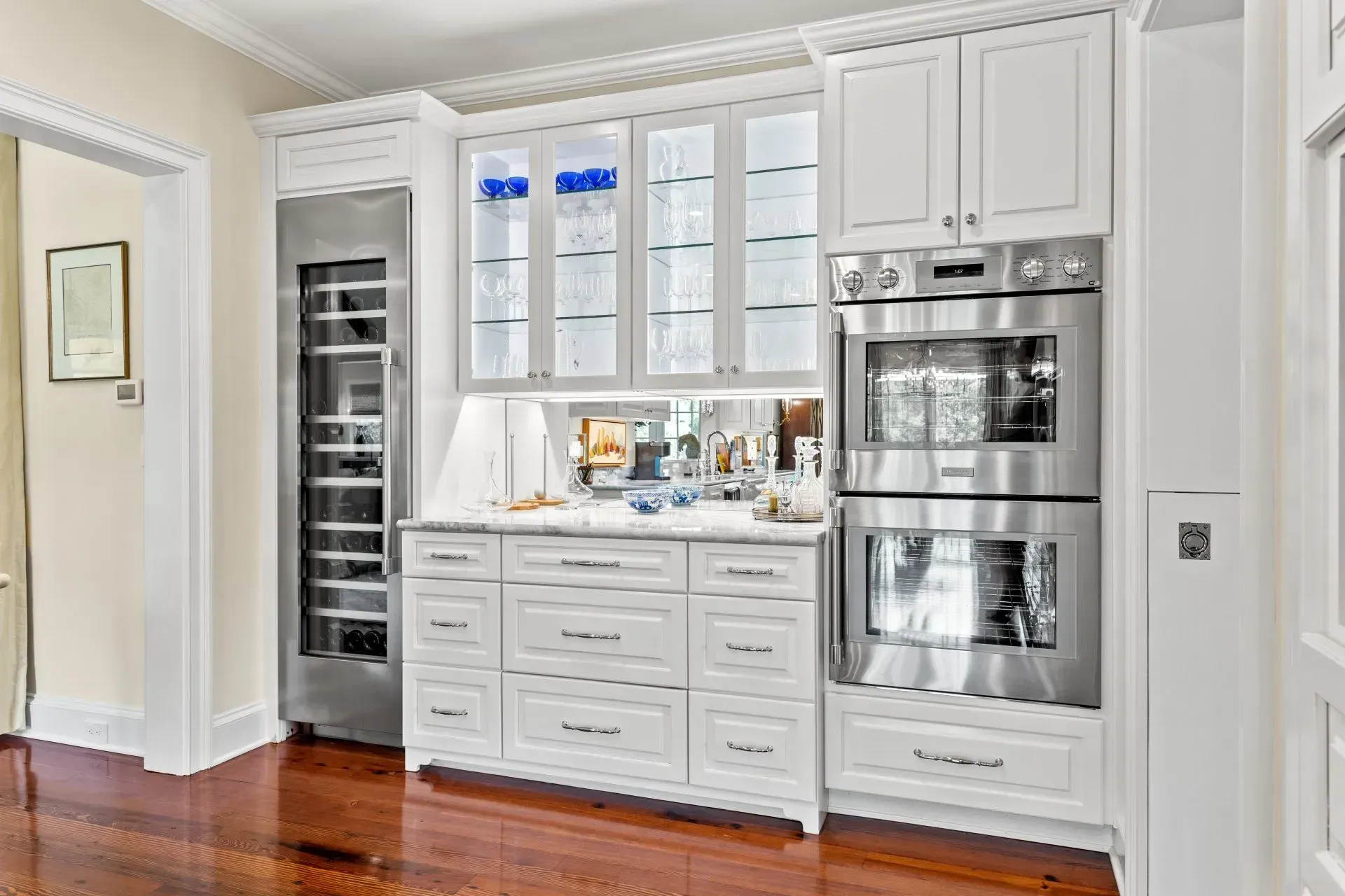 Built-in white kitchen cabinetry with appliances including a wine fridge, oven, and glass-fronted display cabinets.