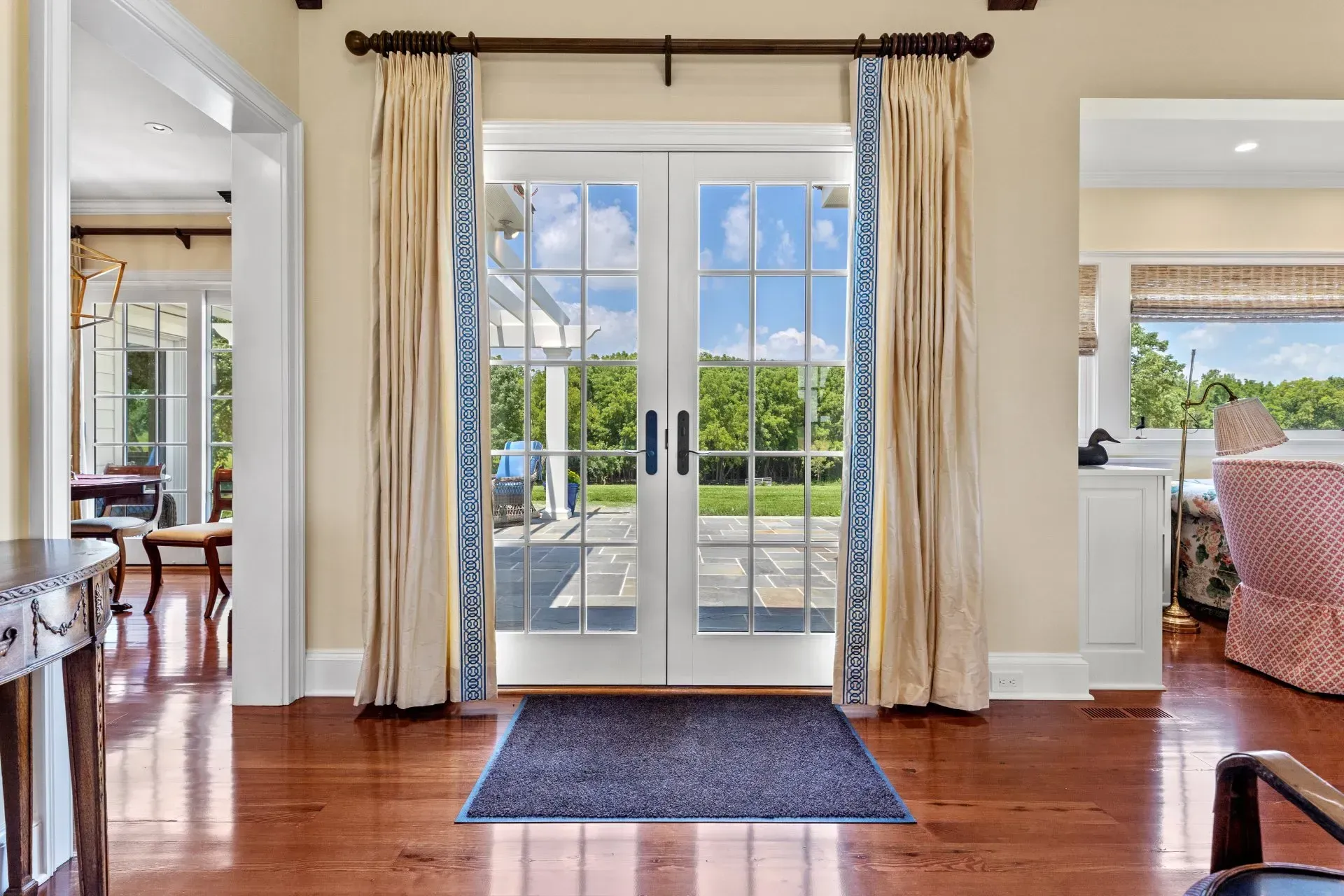 French doors with blue-trimmed curtains open to a yard, with a wooden floor, rug, and cream walls.