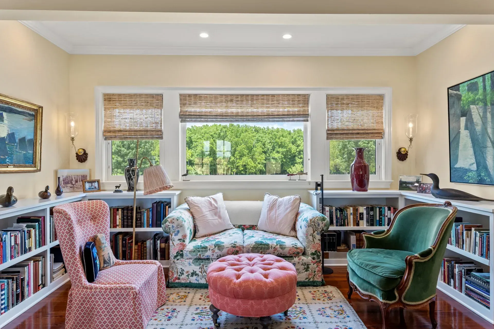 Cozy reading nook with floral sofa, pink chair, ottoman, built-in bookshelves, and large window overlooking trees.