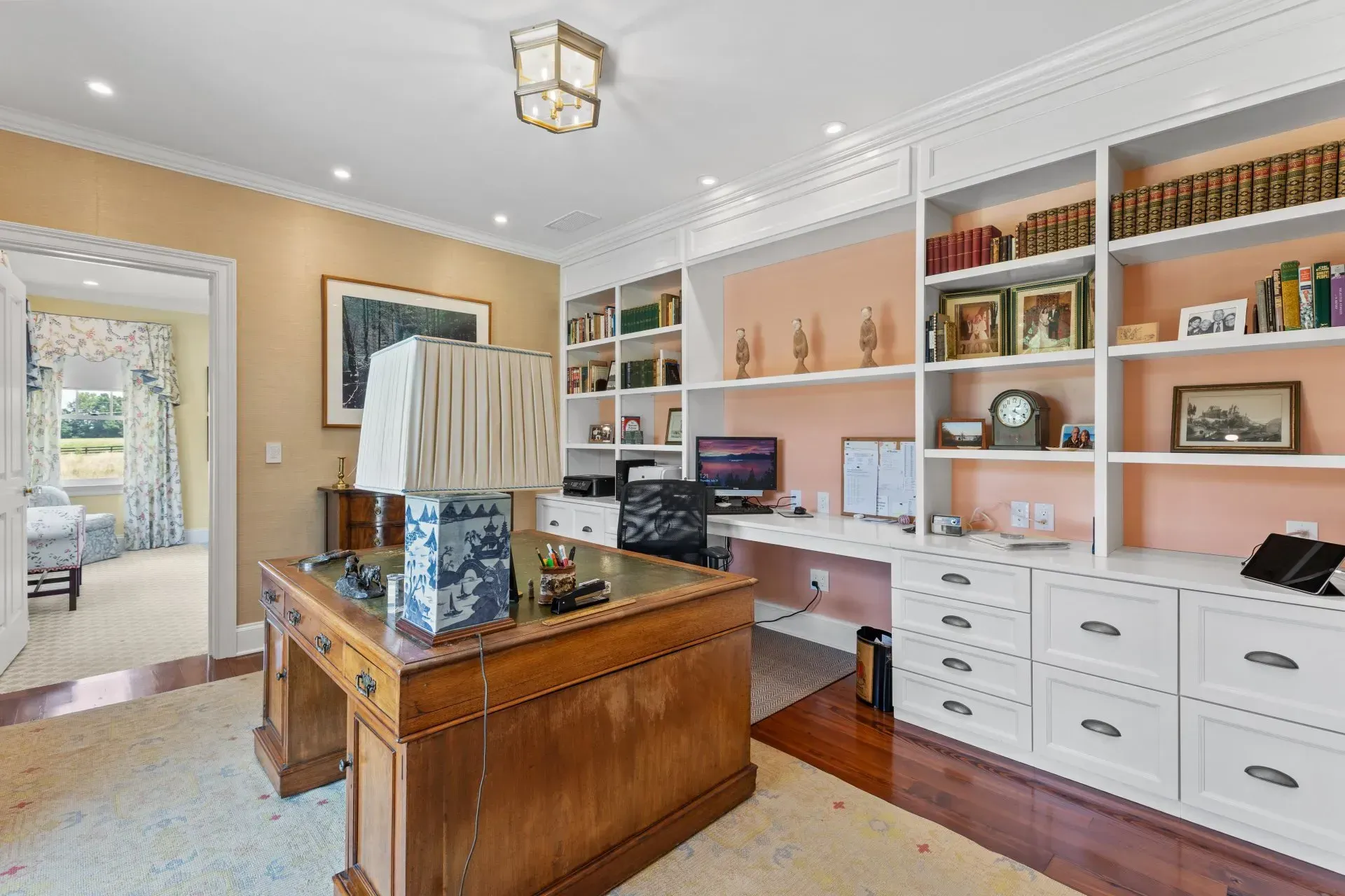 Elegant home office with built-in white shelves, a large wooden desk, and a view into a bedroom.