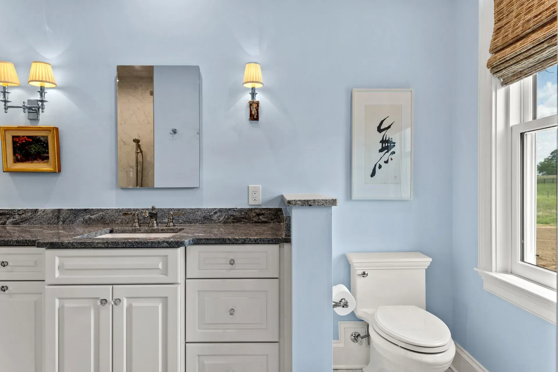 Light blue bathroom with white cabinets, toilet, and a window looking out to a grassy field.