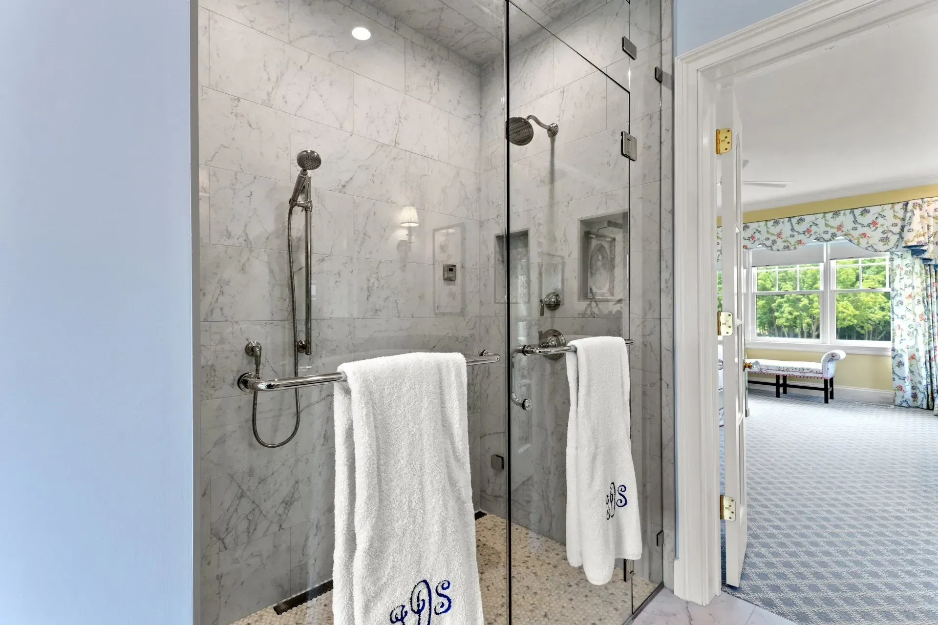 Elegant marble shower with glass doors, white towels. Brightly lit bathroom, doorway to bedroom.