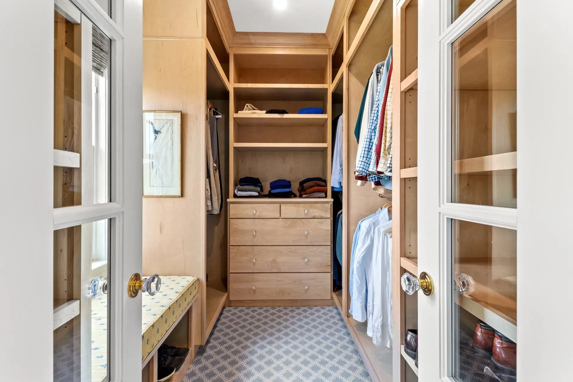 Walk-in closet with wooden shelves, drawers, and hanging clothes. Two doors lead into the room.