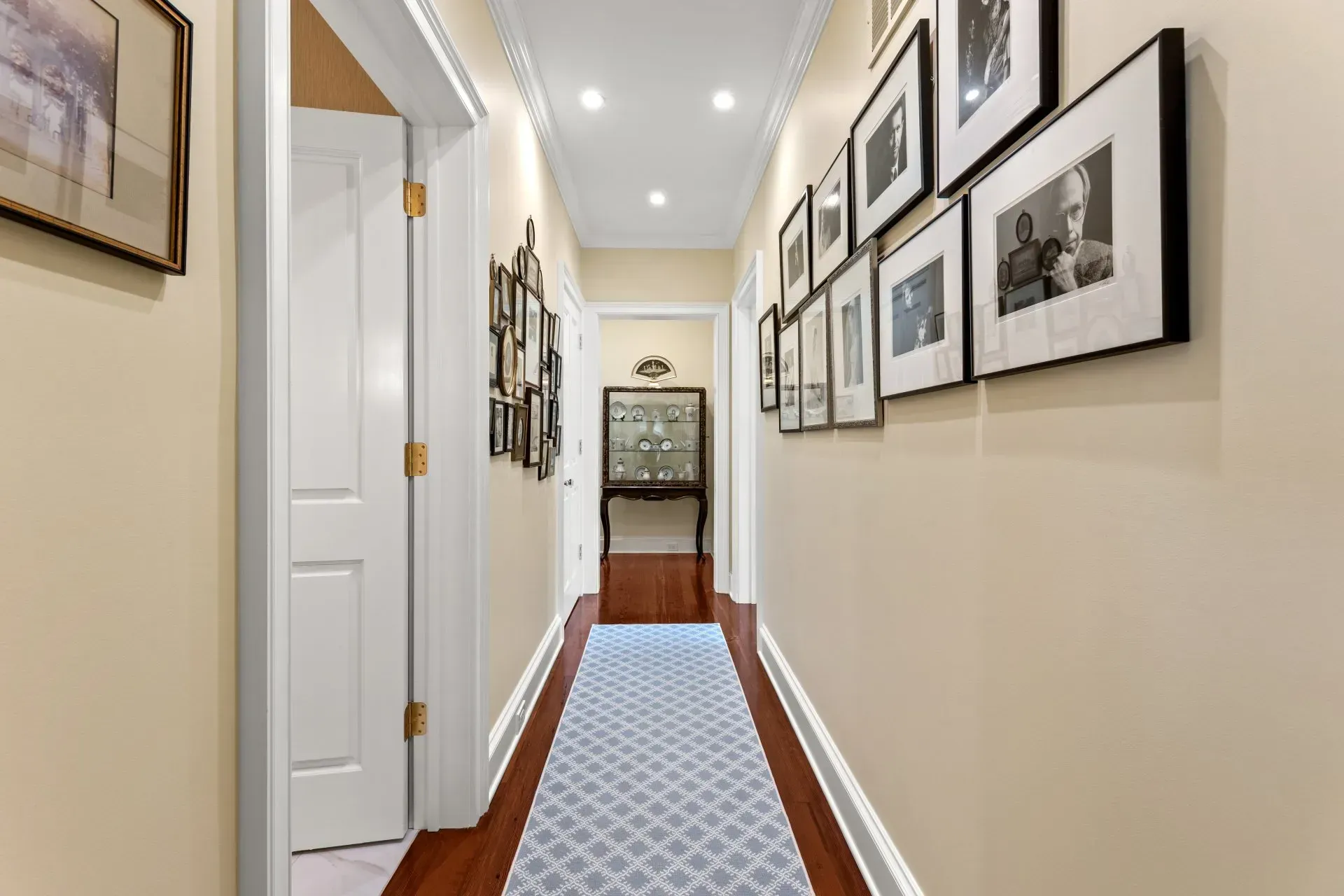 Narrow hallway with framed photos and doors. Light walls, dark wood floors, and a blue runner.
