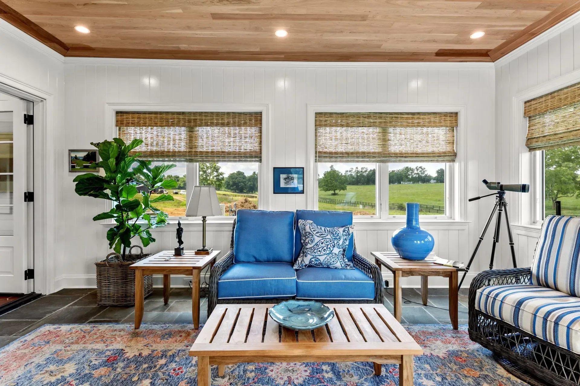 Sunroom with blue furniture, wood ceiling, and view of a grassy field.