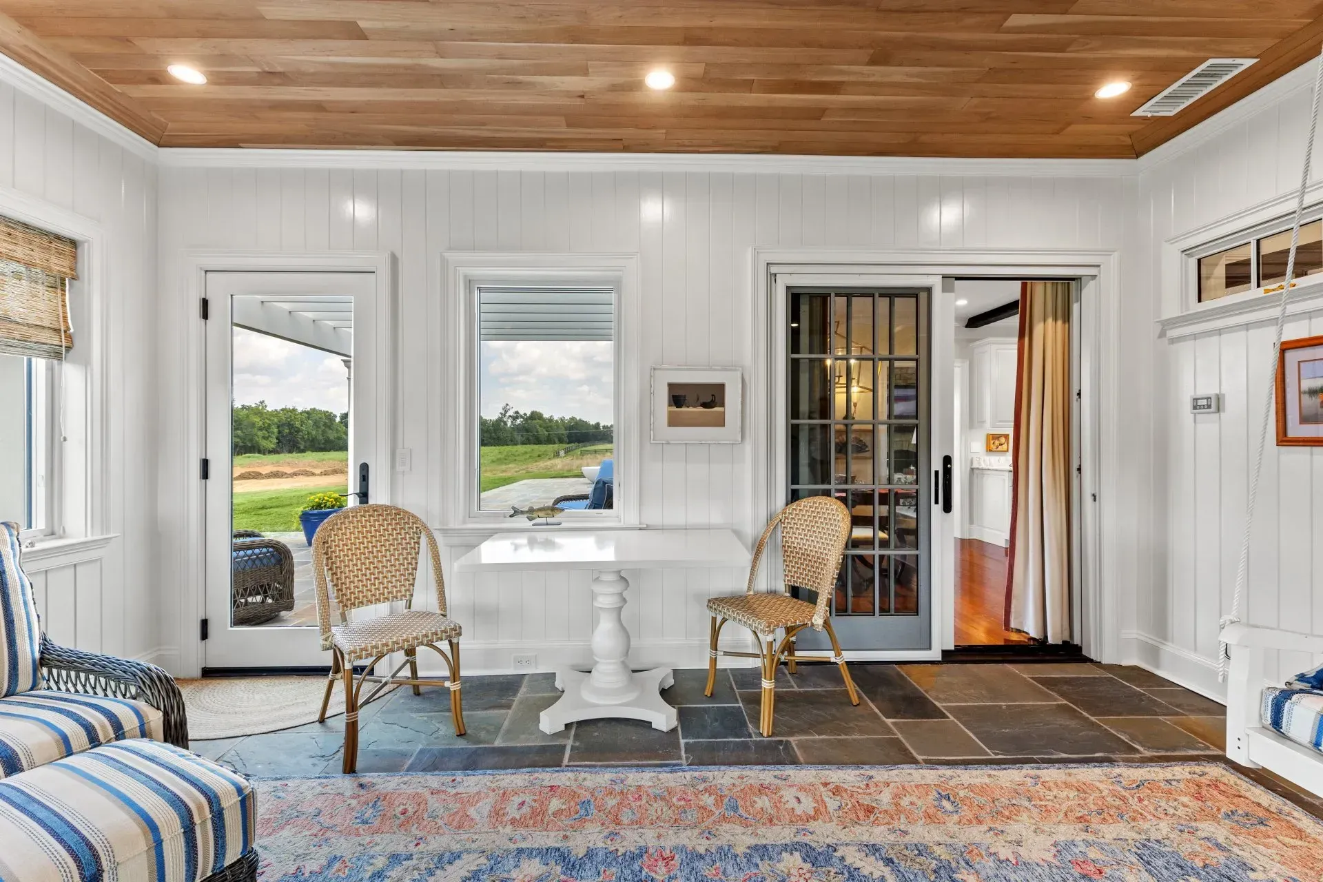 Sunroom with white paneling, wood ceiling, and wicker furniture overlooking a field.