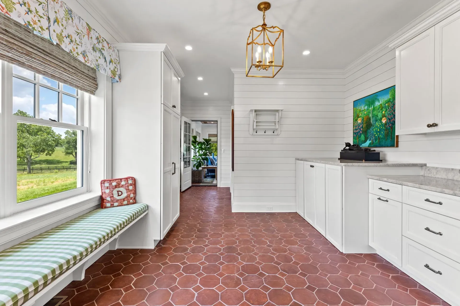Bright laundry room with terracotta tile, white cabinets, and a window seat overlooking a green field.