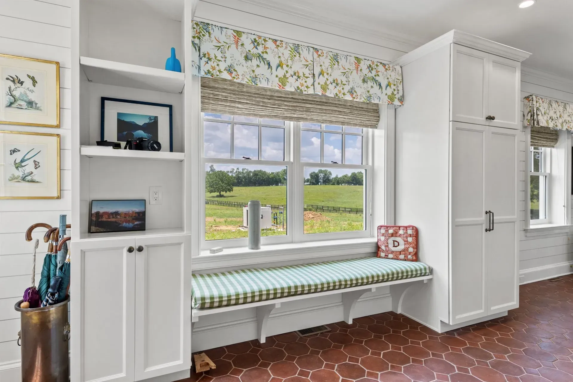 Built-in white cabinets surround a window with a green-cushioned bench, overlooking a grassy field.