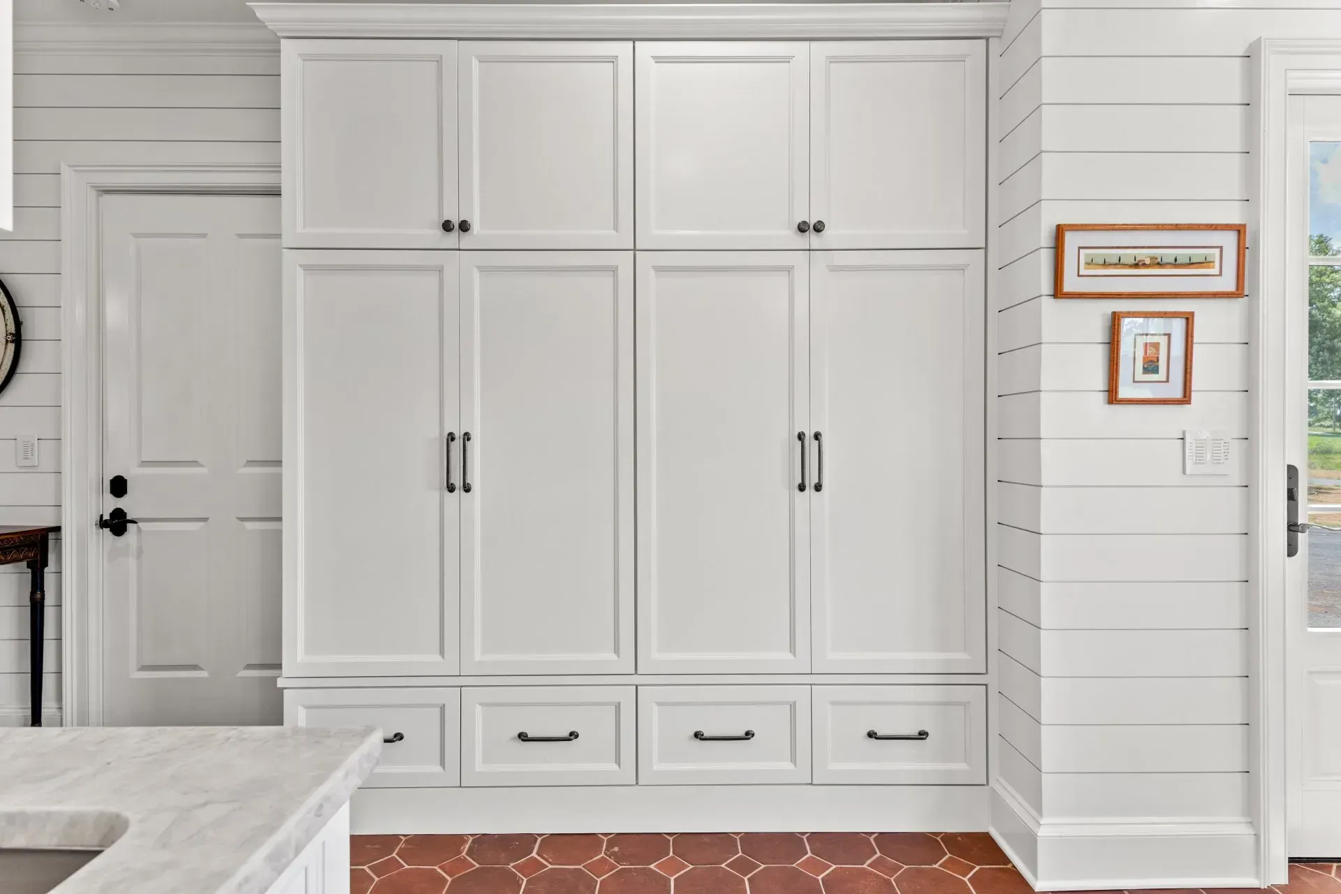 White kitchen pantry with upper cabinets, lower drawers, and black hardware, set against white shiplap walls, on a terracotta tile floor.