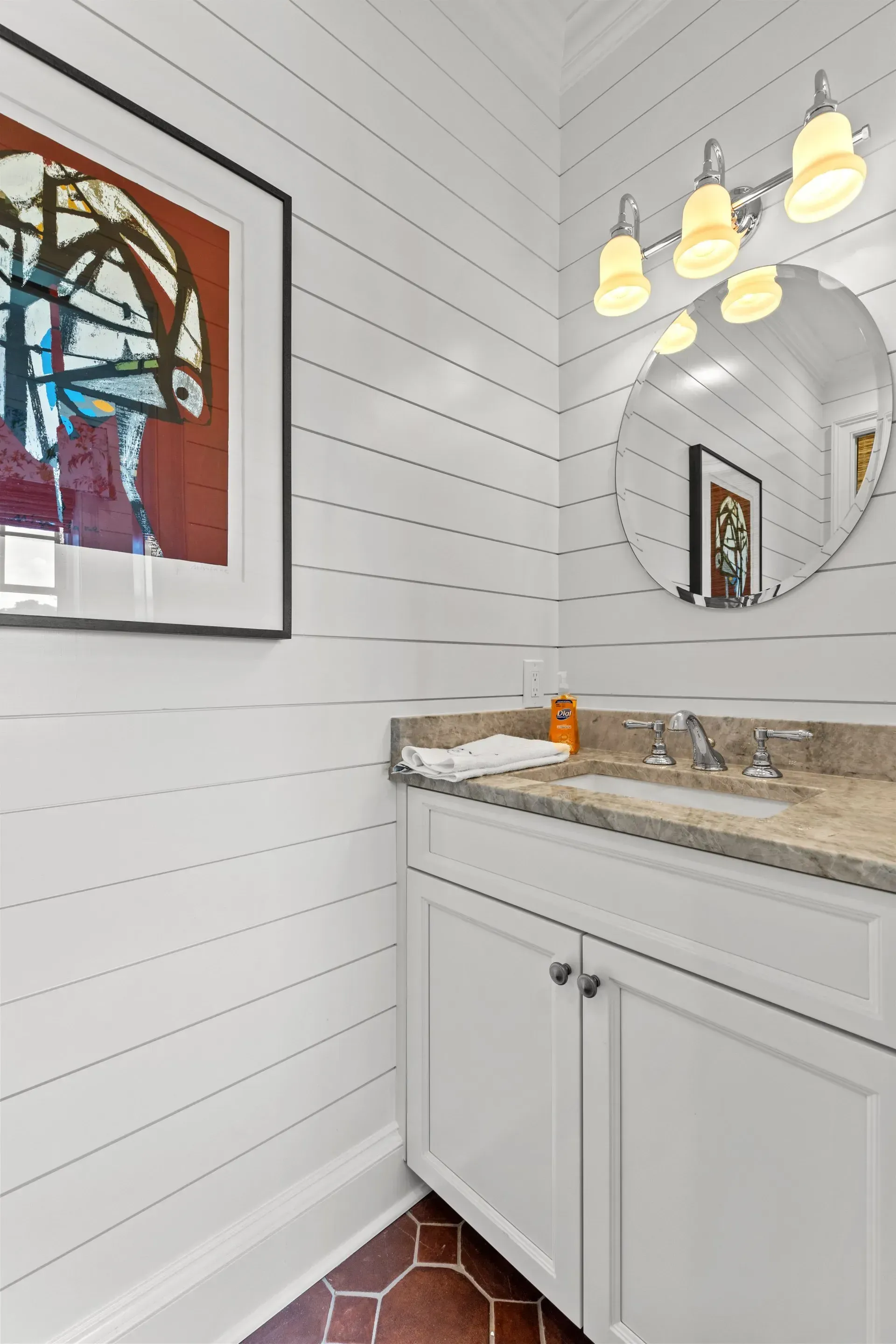 White bathroom with shiplap walls, round mirror, white vanity, and art.