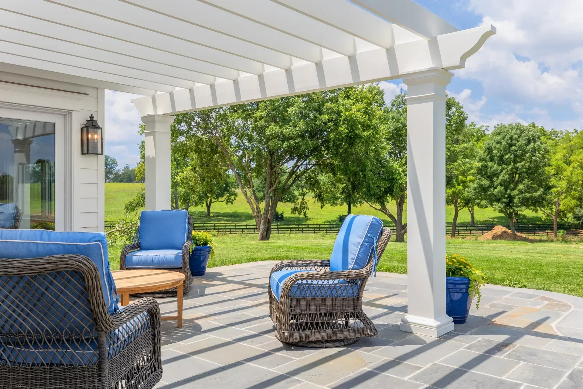 Patio with white pergola, blue chairs, and a wooden table. Green grass and trees in the background.