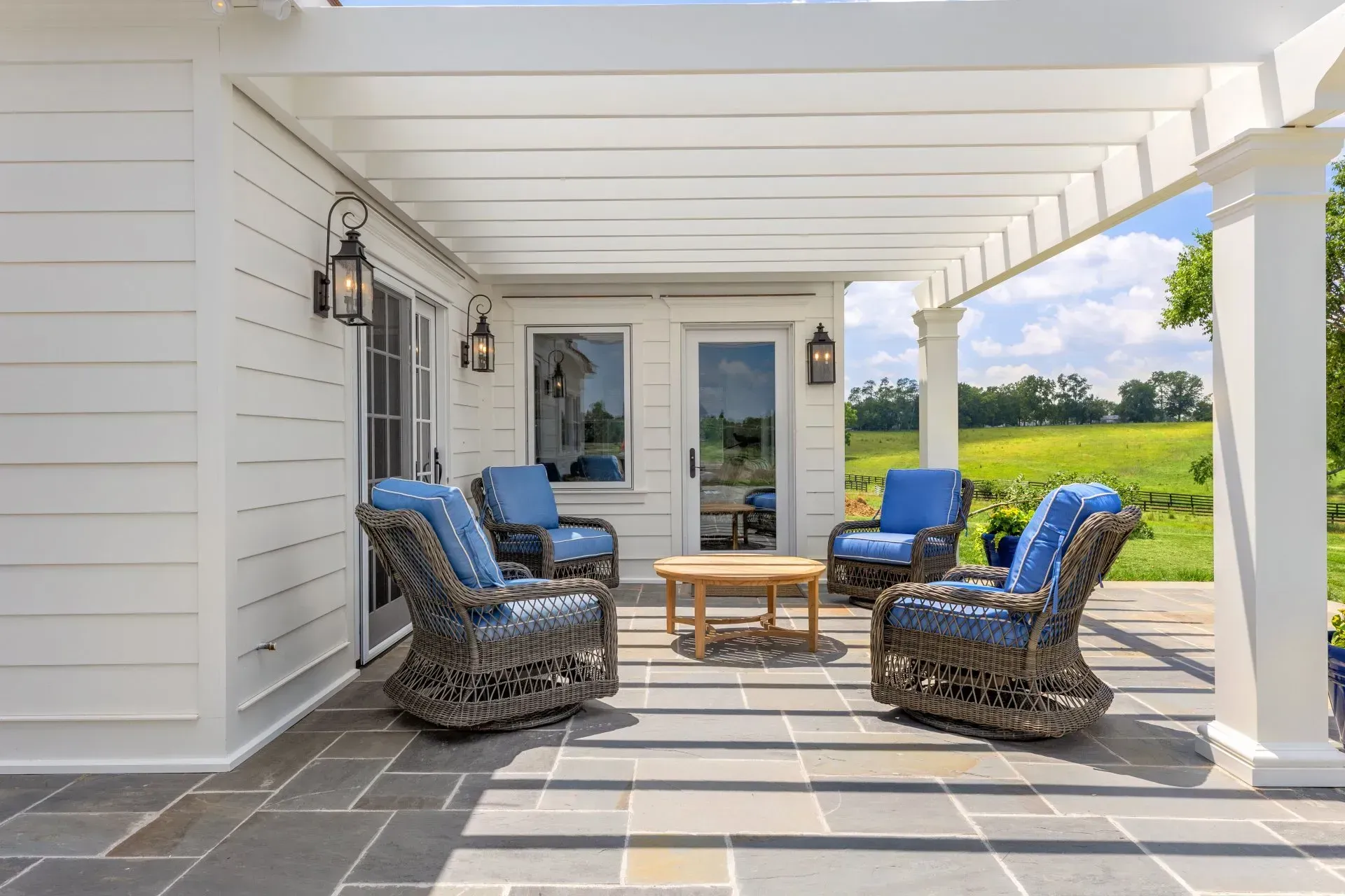 Patio with blue chairs, wooden table, and white pergola overlooking a field.