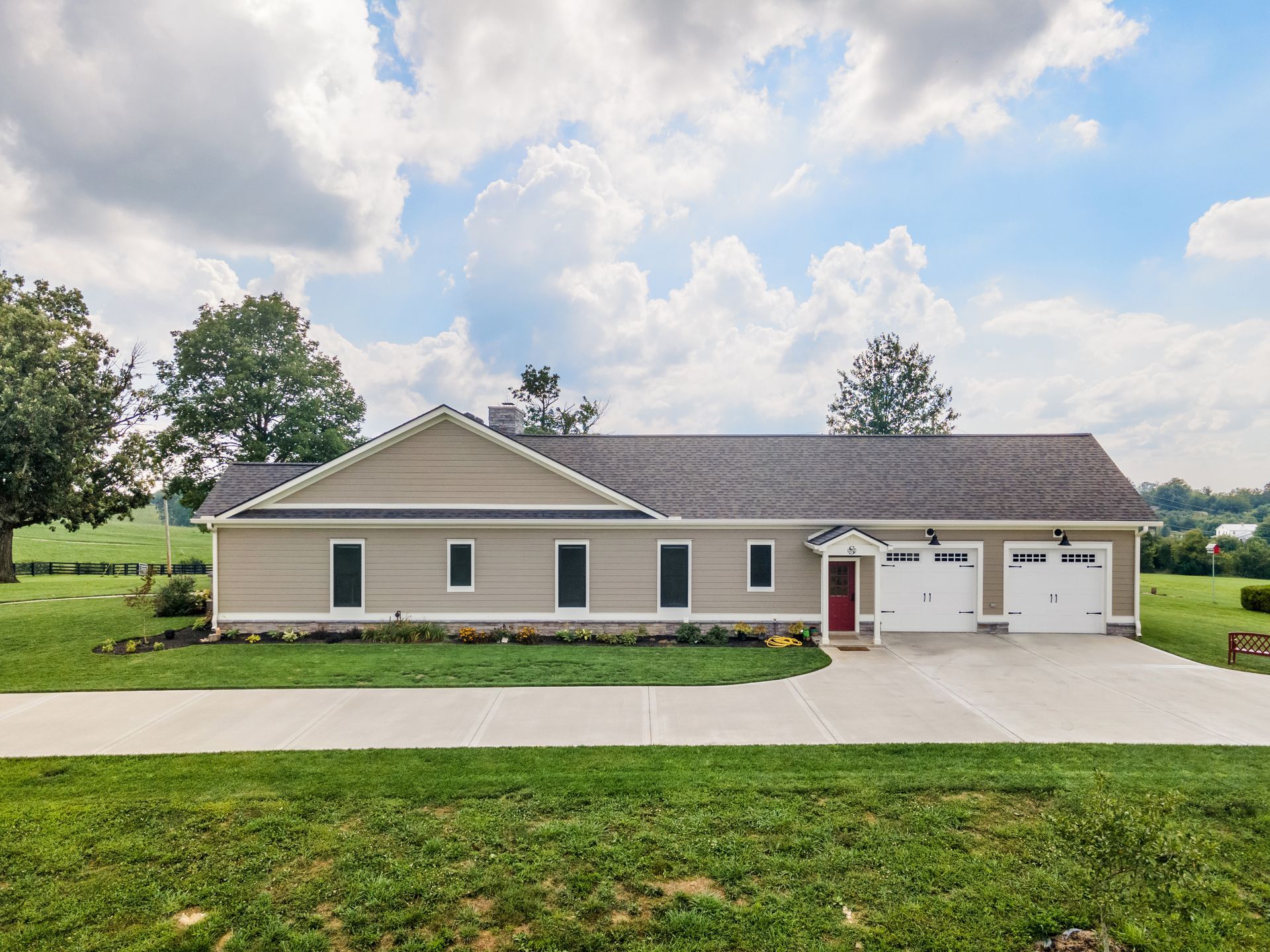 Tan ranch house with three-car garage, red front door, and long driveway on a grassy lawn under a cloudy sky.