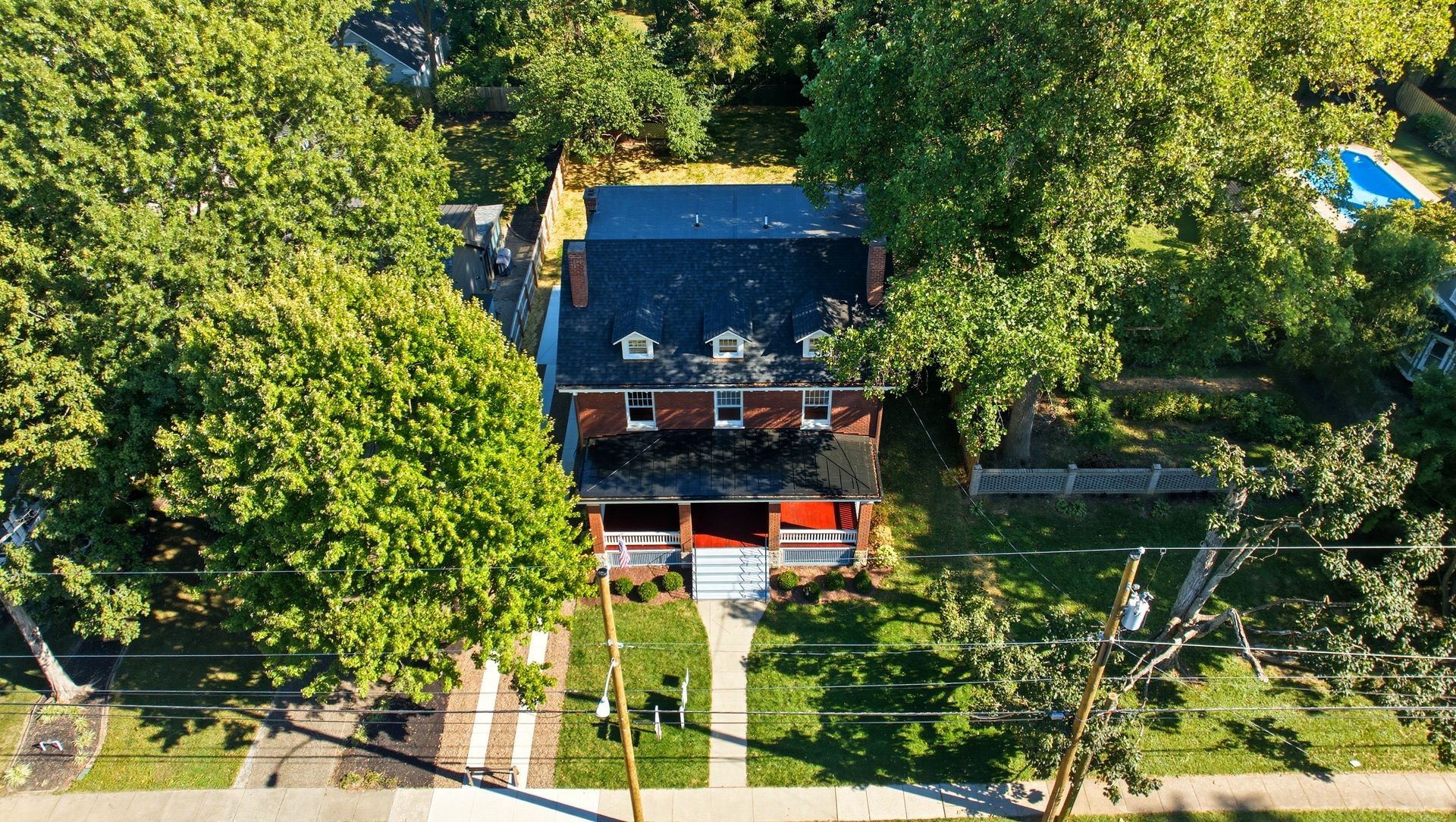 Red brick house with a porch and front yard, surrounded by green trees, aerial view.