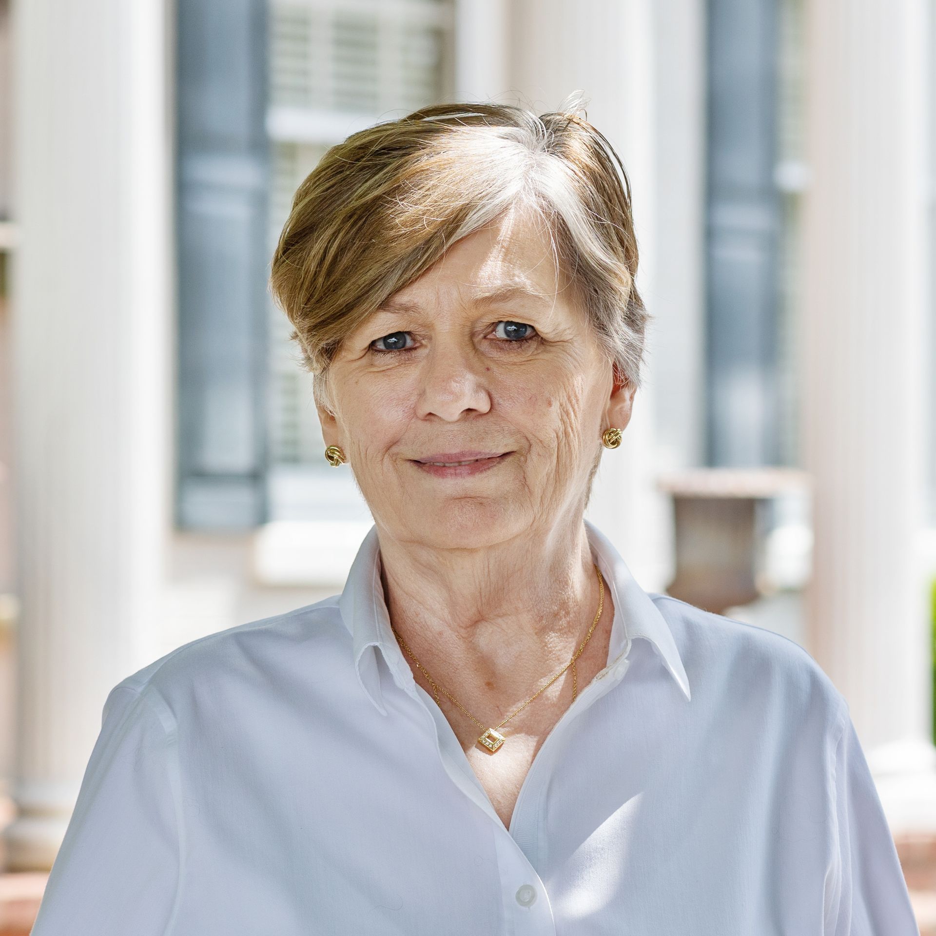 Woman with short blonde hair and a white shirt smiles outdoors near a building with columns.