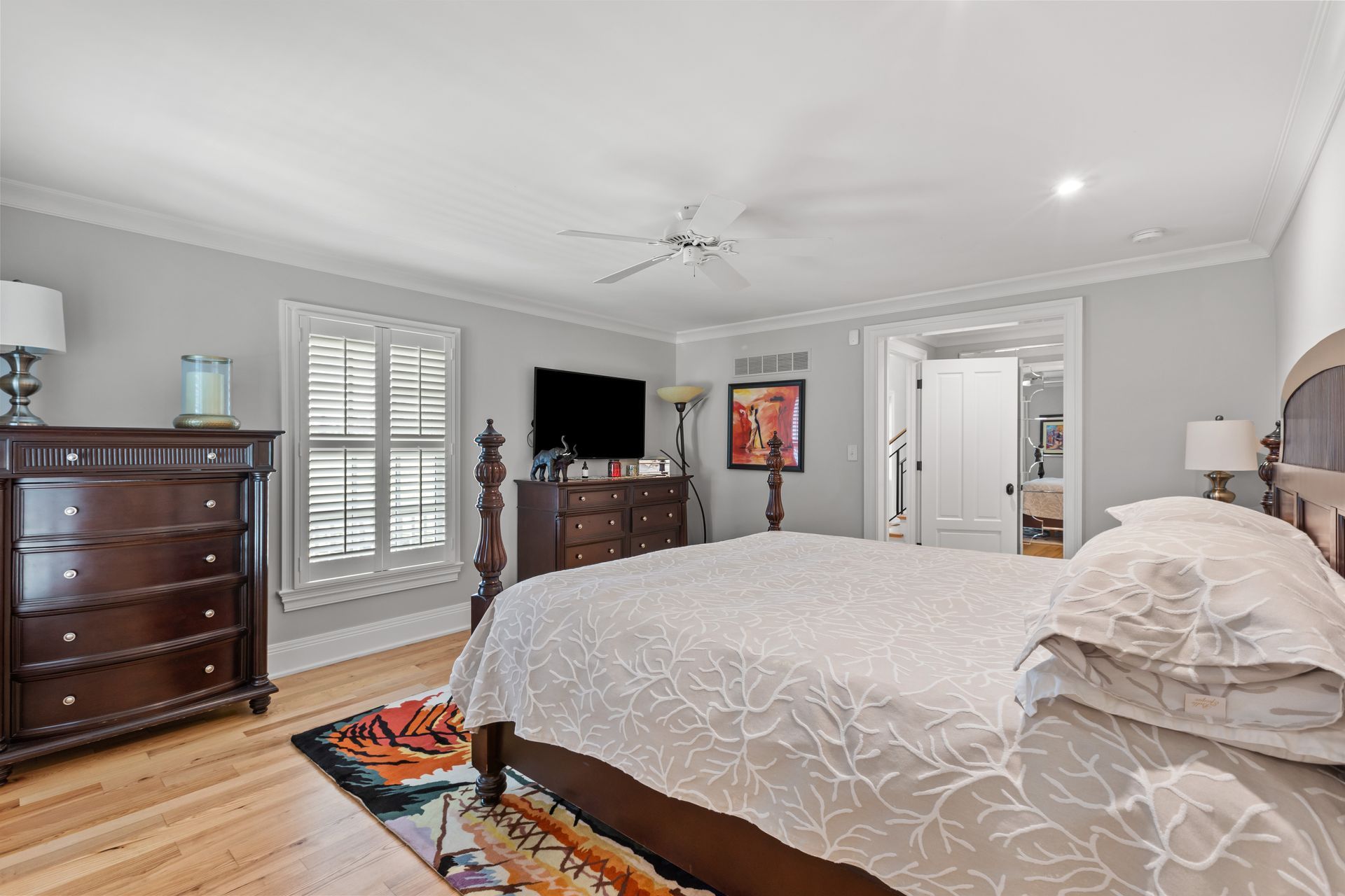 Bedroom with bed, dresser, TV, shutters, hardwood floor, and a doorway to a bathroom.