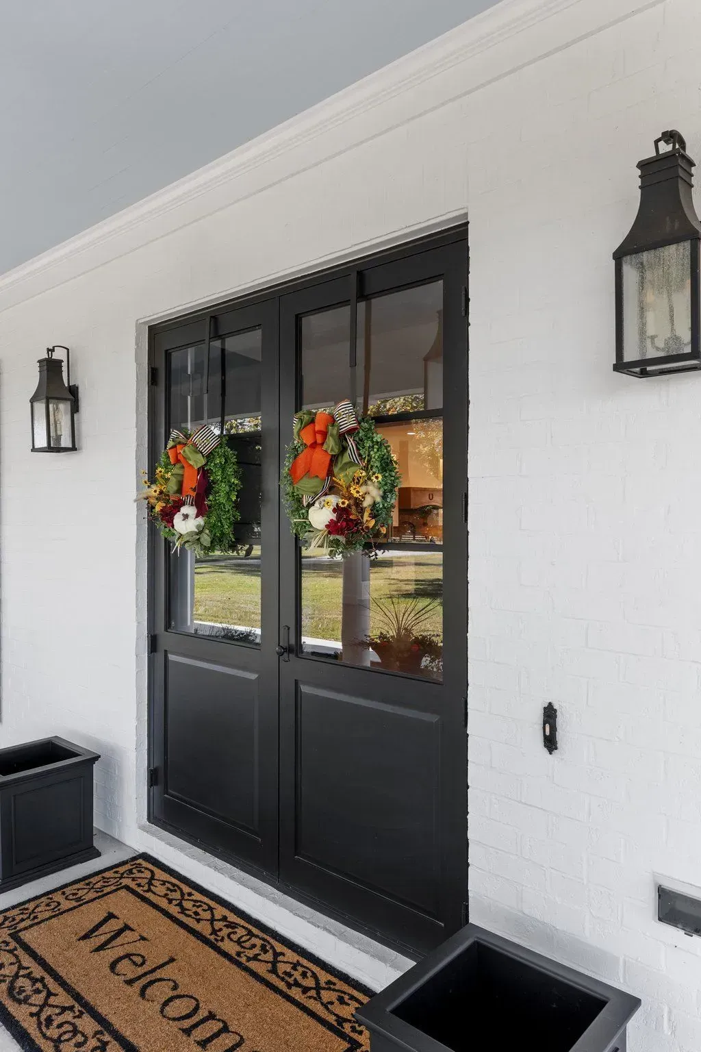 Black double doors with wreaths and matching planters on a white brick wall.