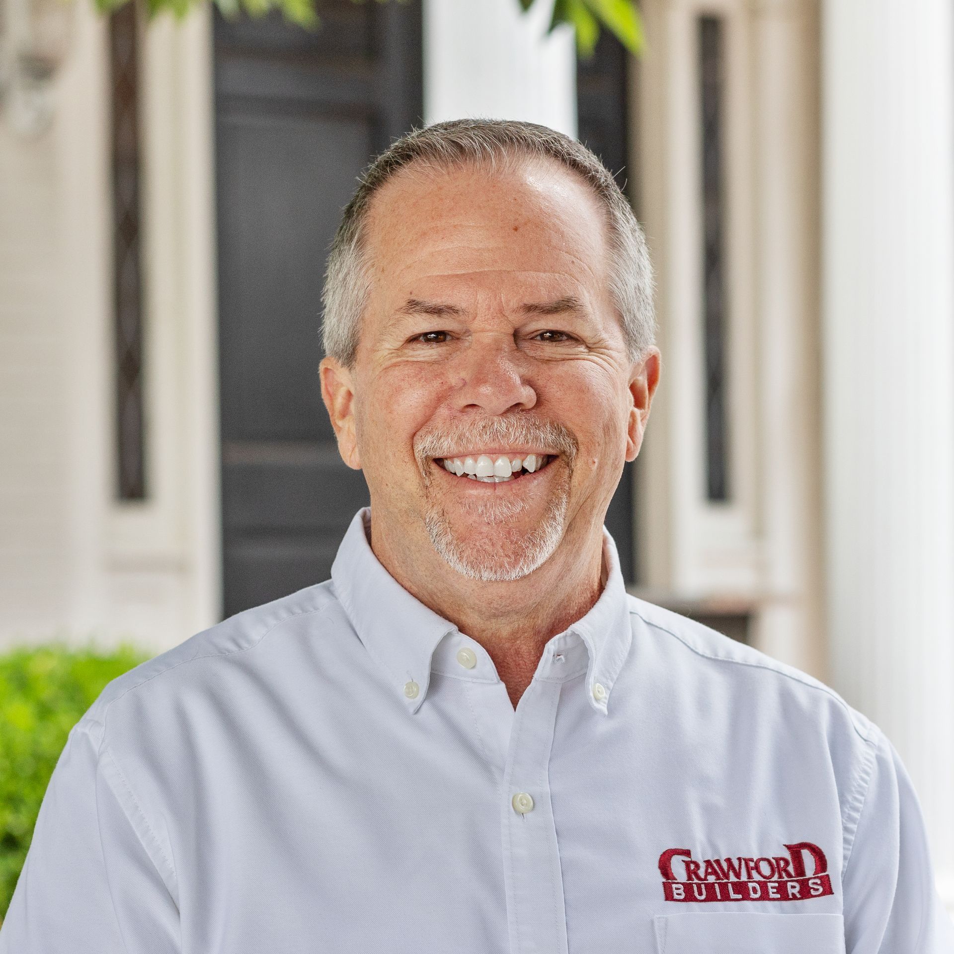 Smiling man with graying hair wearing a white shirt, standing outdoors near a building.