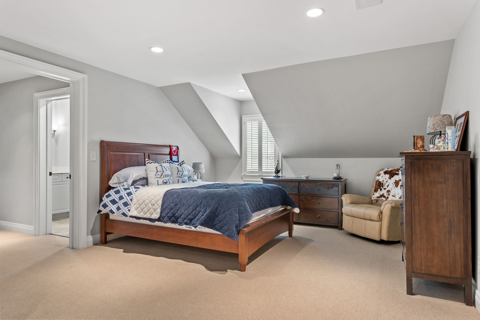 Bedroom with a bed, dresser, armchair, and a doorway to a bathroom; beige carpet, gray walls.