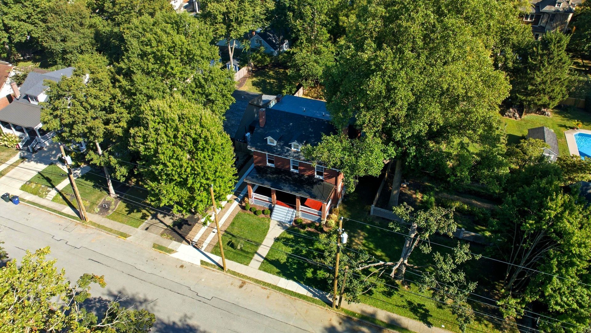Aerial view of a brick house surrounded by lush green trees, a pool, and a street on a sunny day.