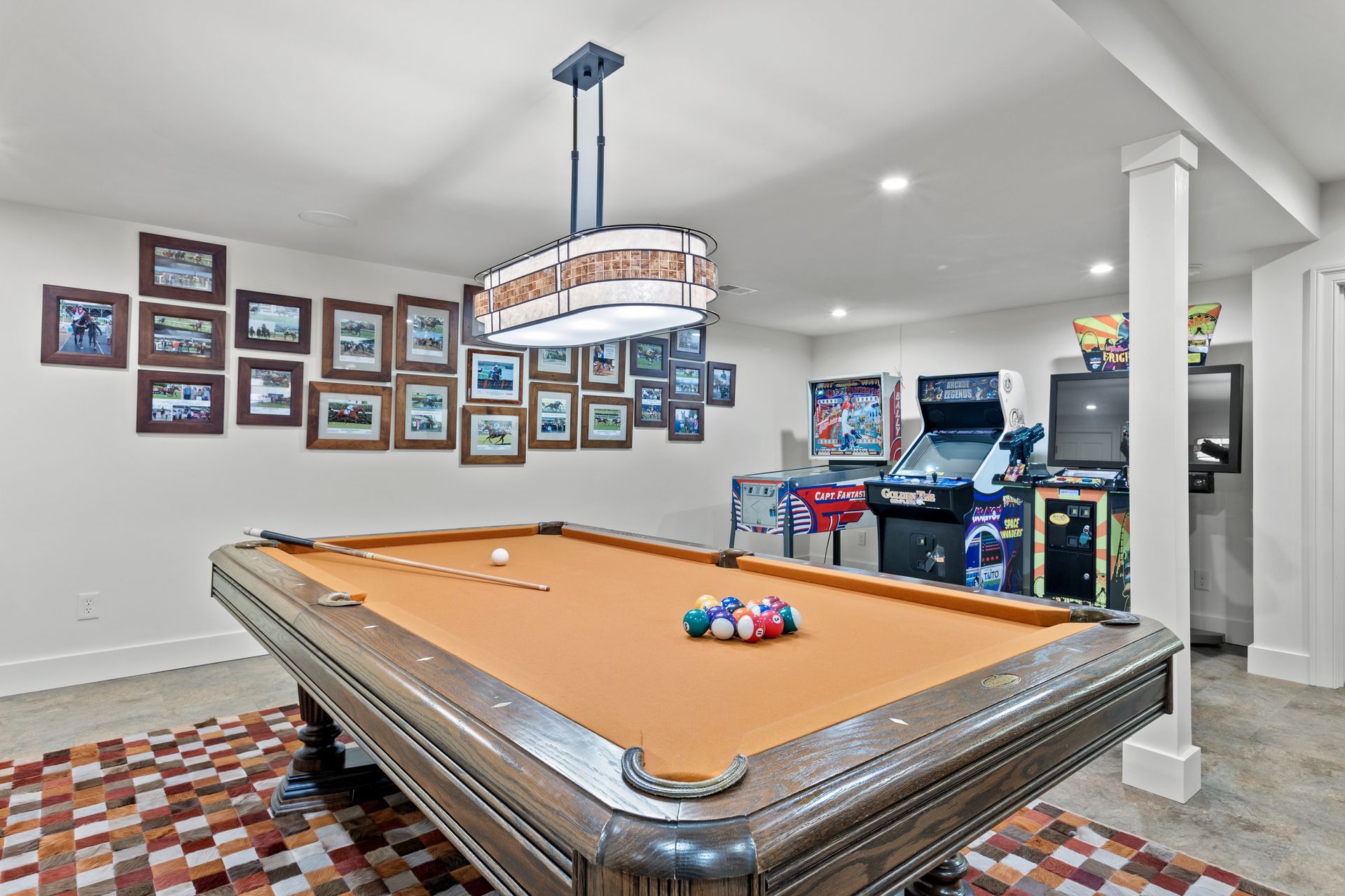 Pool table in a rec room with arcade games, framed photos, and patterned rug.