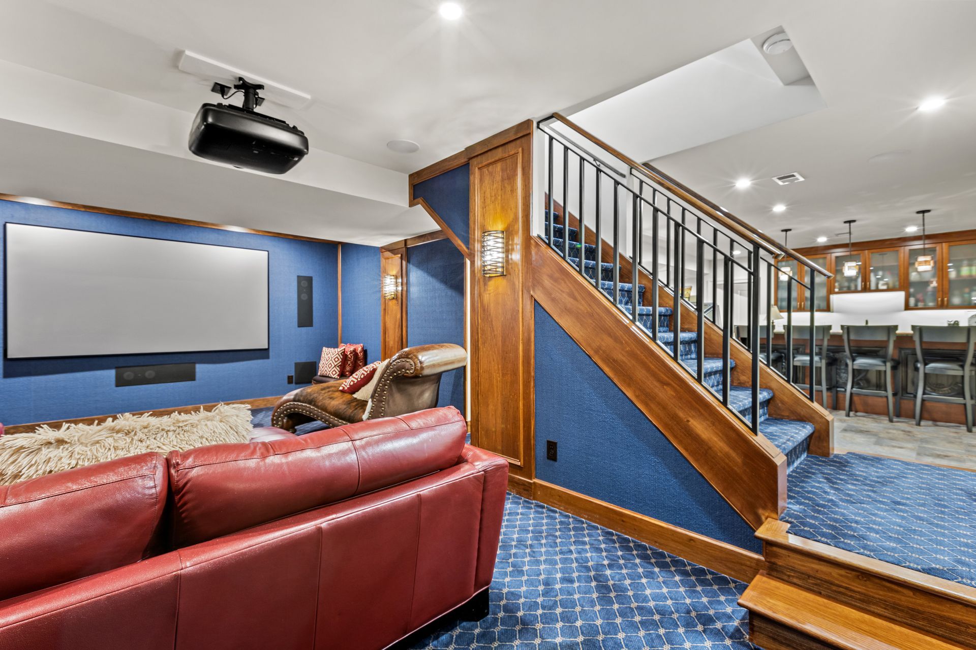 Basement home theater with blue walls, red couch, projector, bar area, and wooden staircase.