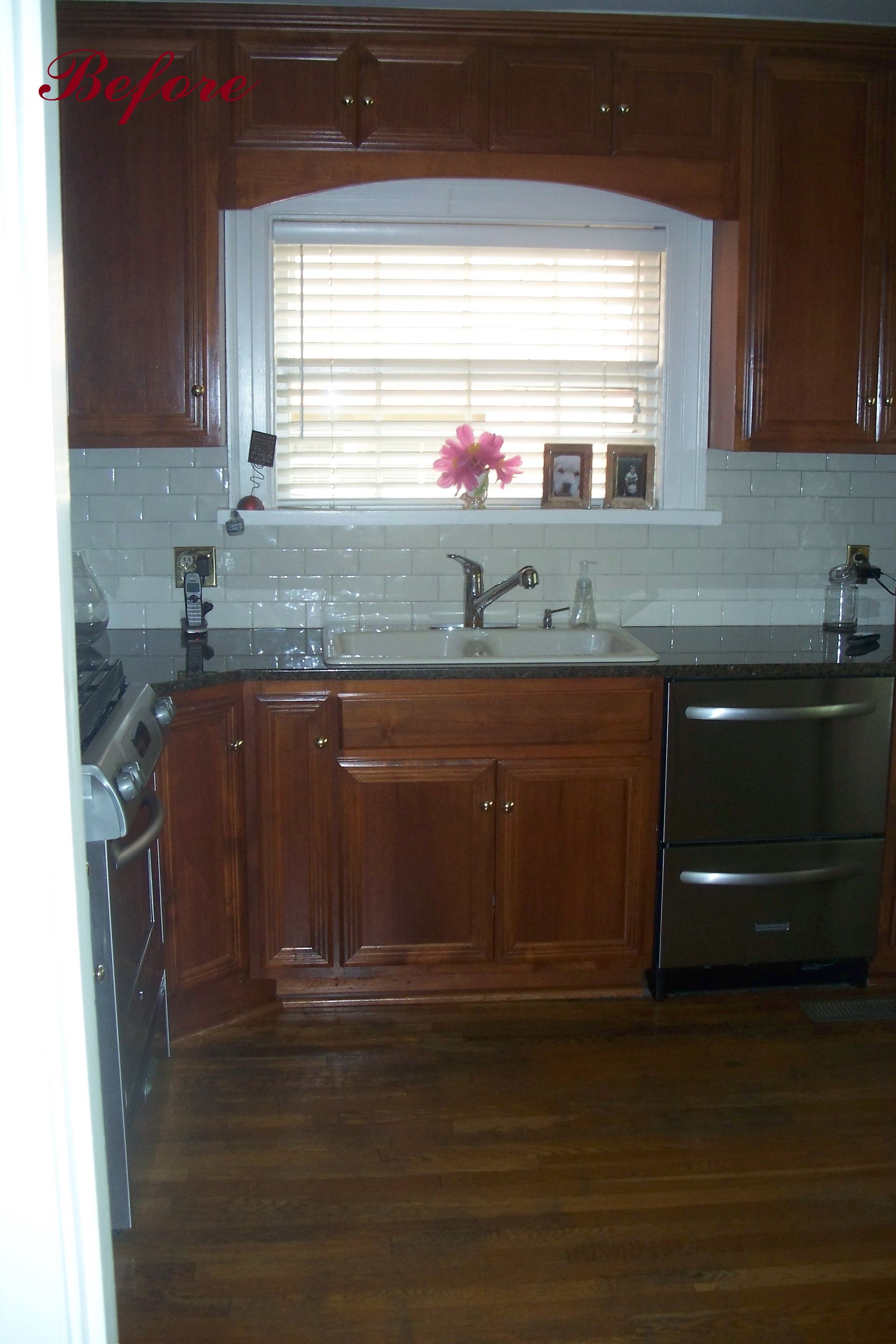 Kitchen with wood cabinets, dark countertops, and stainless steel dishwasher. Window with blinds, and flowers on the sill.