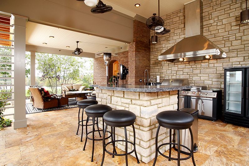 Outdoor kitchen with stone facade, granite countertop, and stools. Grill, fridge, and covered seating area.