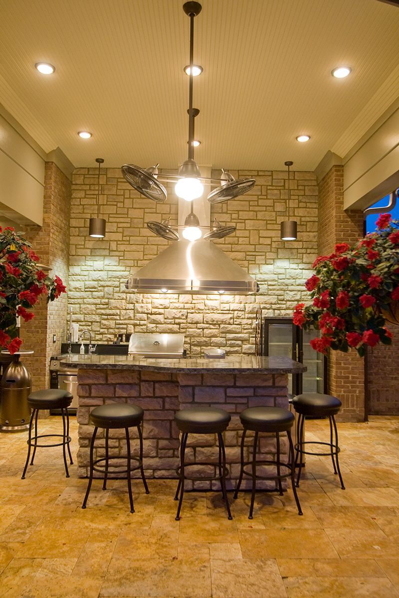 Outdoor kitchen with stone bar, stools, grill, hood, and ceiling fan; roses frame the space.