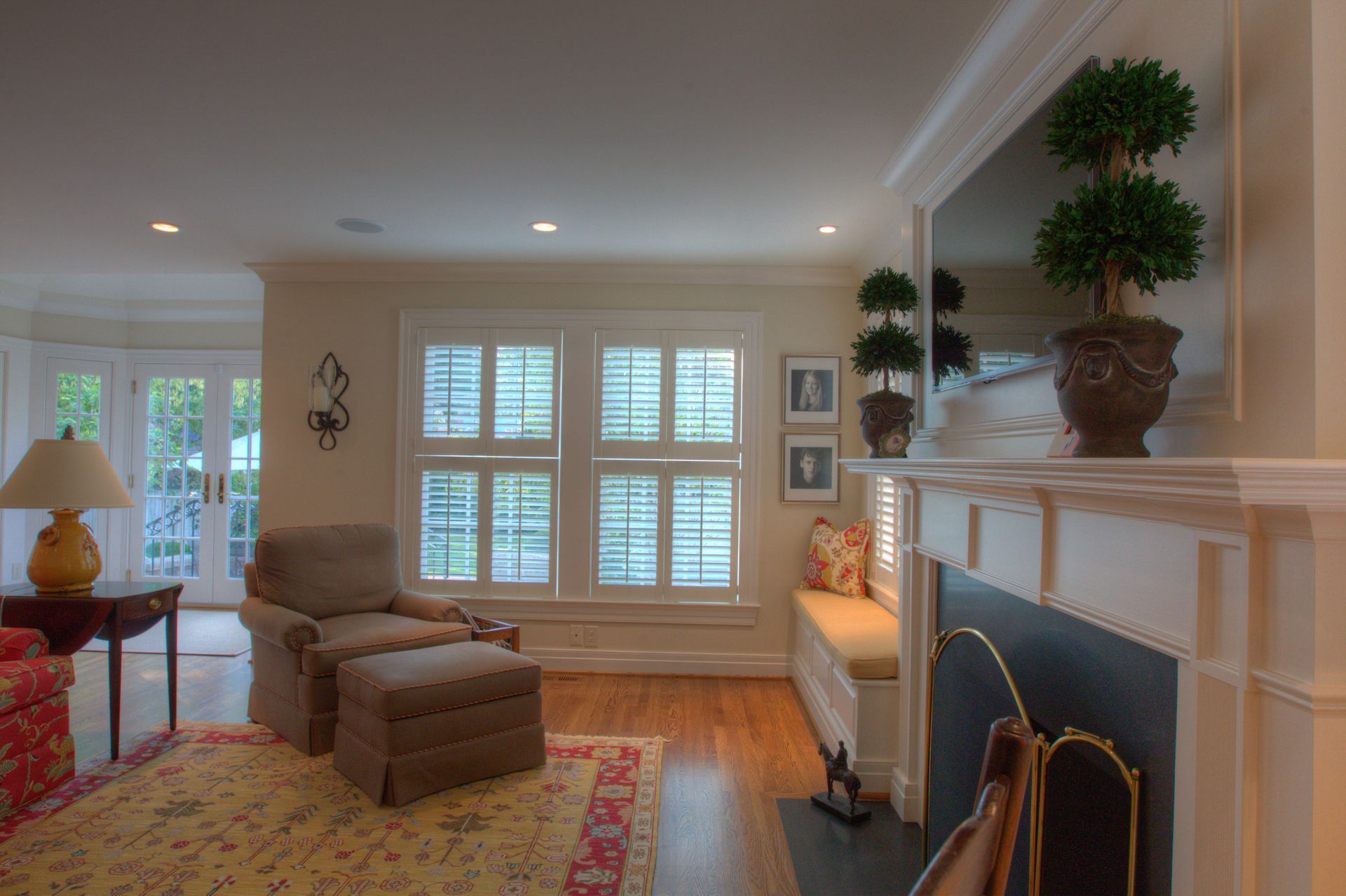 Living room with fireplace, armchair, window seat, and shutters. Soft lighting, neutral colors, wood floors, and a rug.