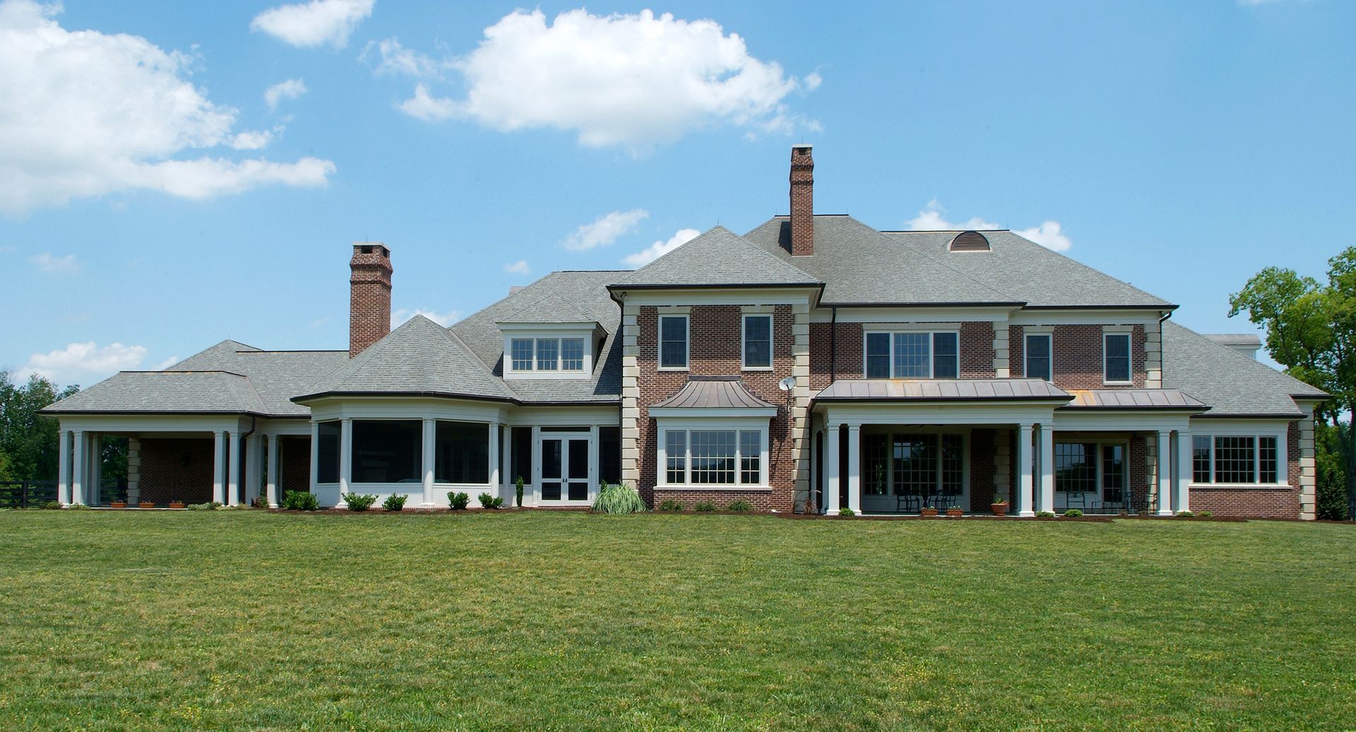 Large, multi-level brick mansion with a gray roof and white columns on a grassy hill under a blue sky.