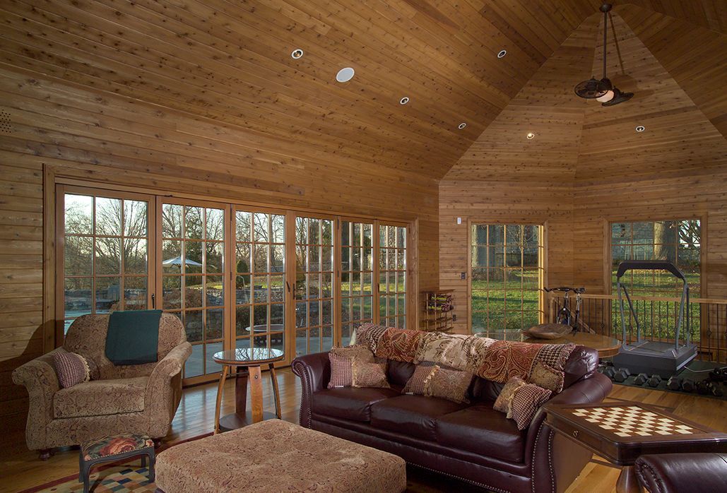 Cozy living room with wooden walls, large windows overlooking a yard, and a leather sofa with blankets and pillows.