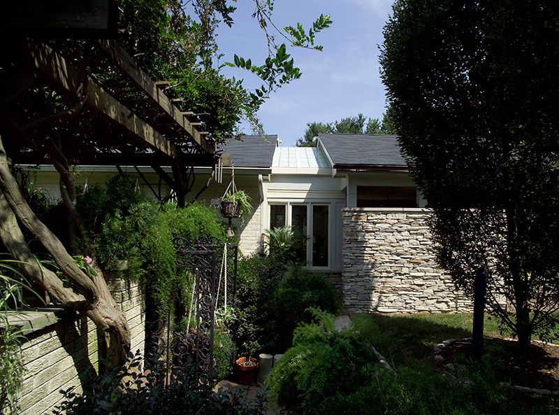 Exterior view of a light-colored house with a stone wall, surrounded by lush greenery and a wooden pergola.