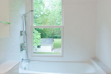 White bathroom with a bathtub, shower, and a window overlooking a yard with a small building and trees.
