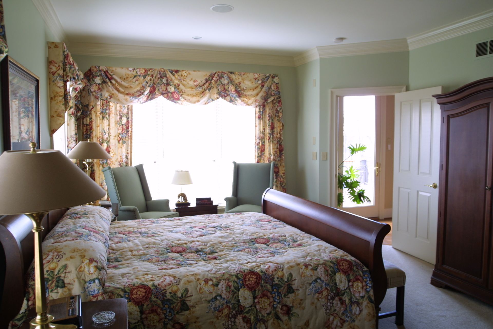 Bedroom with floral bedspread, two chairs by a window with floral curtains, and a dark wooden wardrobe.