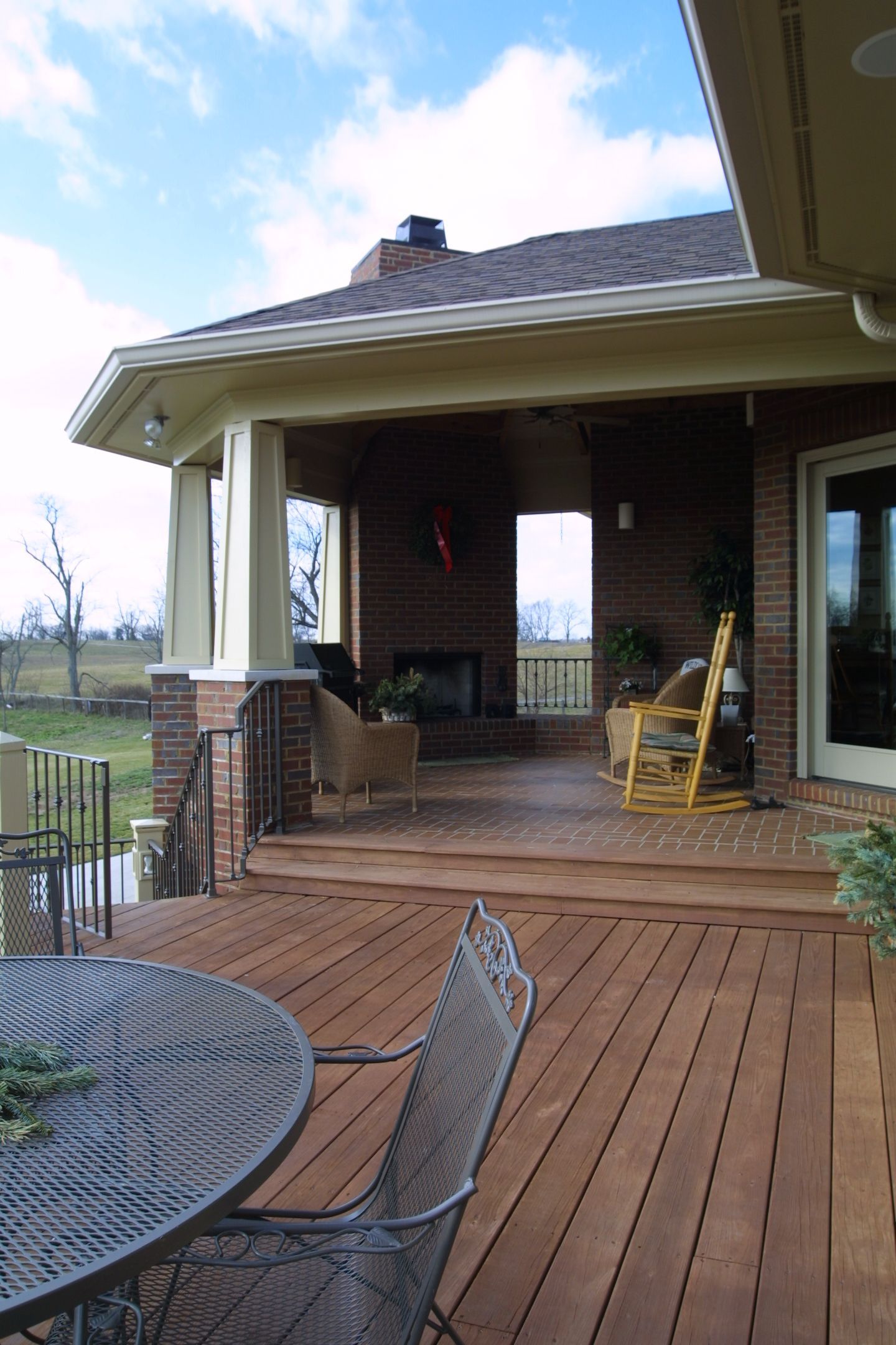 Wooden deck with patio furniture, leading to a covered porch with brick columns and a fireplace.