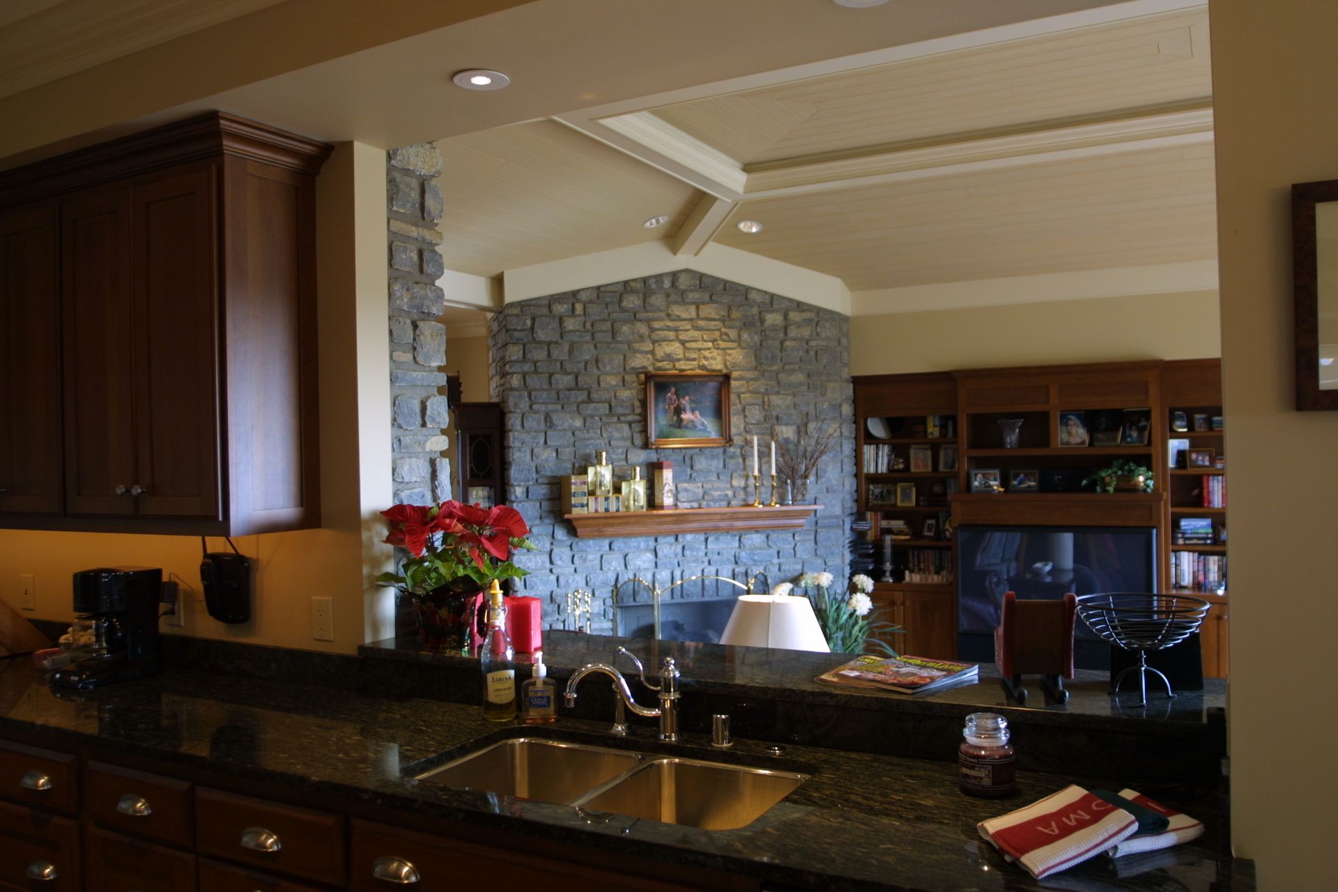 Kitchen with dark cabinets and a sink, looking through a stone-walled opening to a living area with a fireplace and bookshelves.