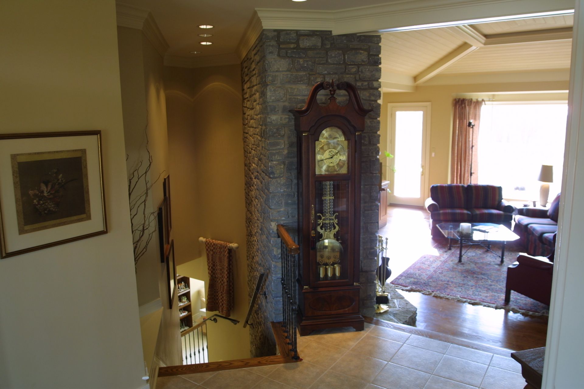 Entryway with staircase, grandfather clock, and view into living room with sofa, rug, and windows.