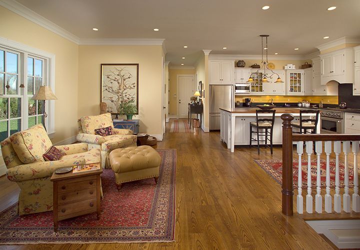 Cozy living room with floral chairs, rug, and kitchen with island; hardwood floors; yellow walls.