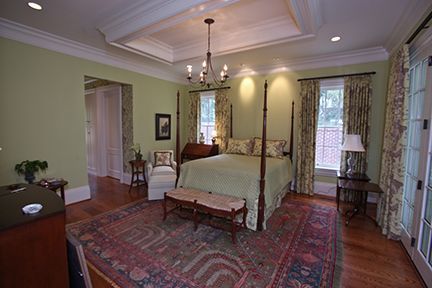 Bedroom with four-poster bed, antique rug, light green walls, and windows with patterned curtains.