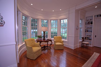 Curved room with hardwood floors, yellow armchairs, table, and windows overlooking a yard.
