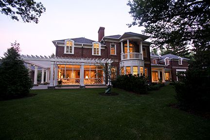 Large brick mansion at dusk, with a grassy lawn and pergola. Lit windows.